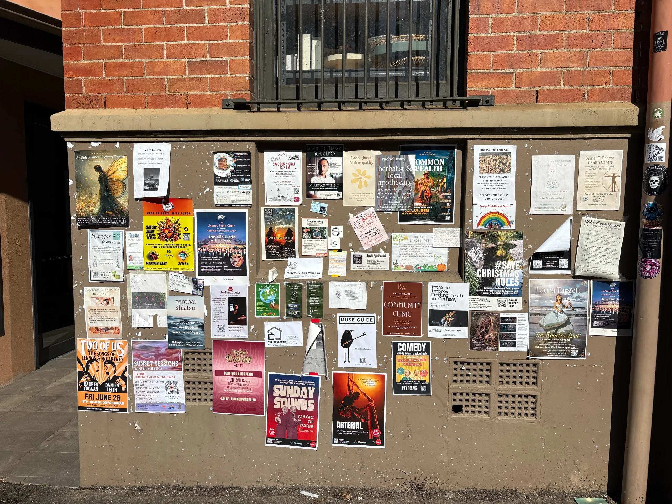 A community noticeboard covered in posters for gigs and markets in Bellingen