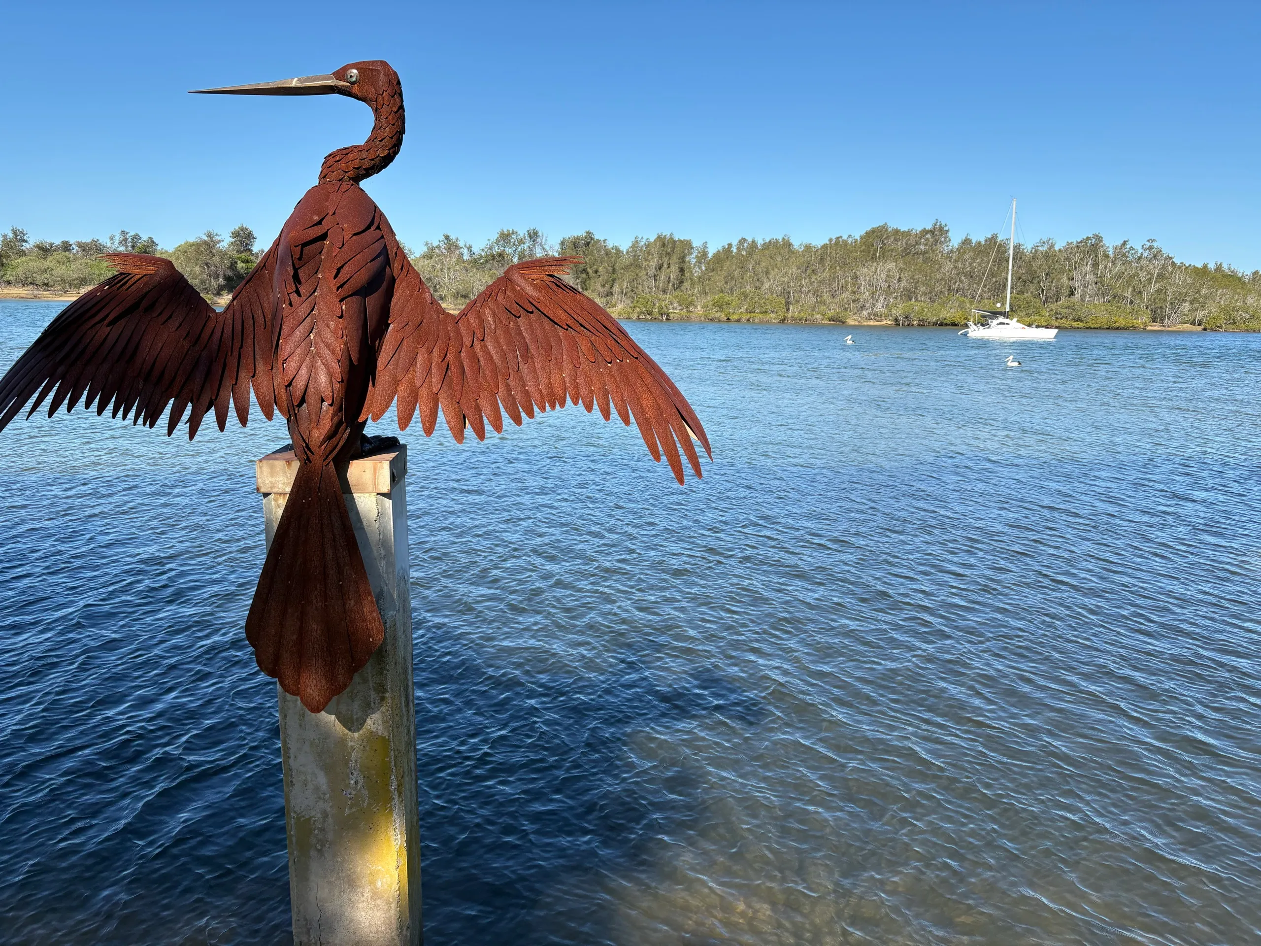 A rusted metal darter sculpture with wings spread on a pylon over the Nambucca River