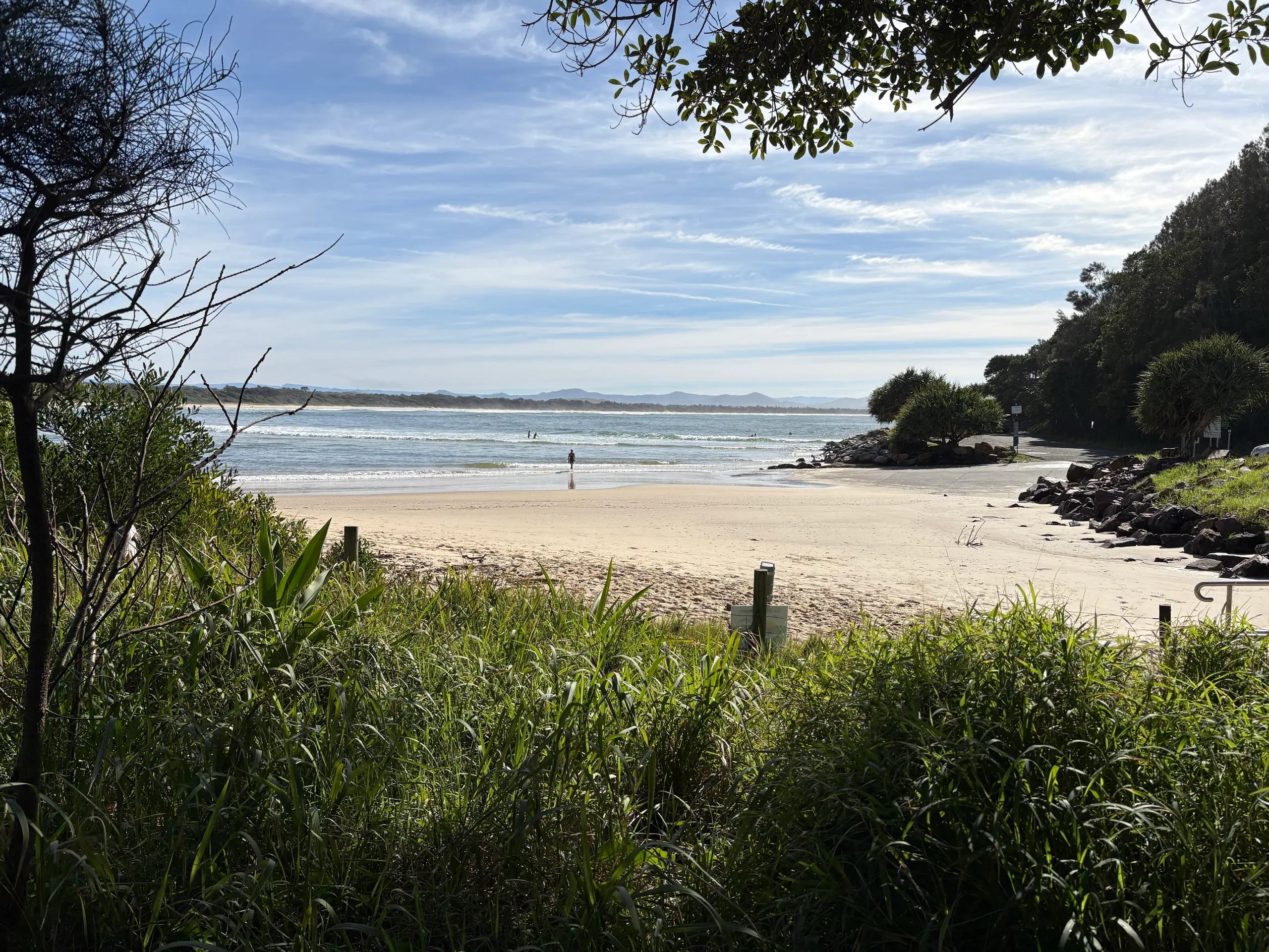Scotts Head Main Beach seen through coastal trees, with the headland and rocks on the right