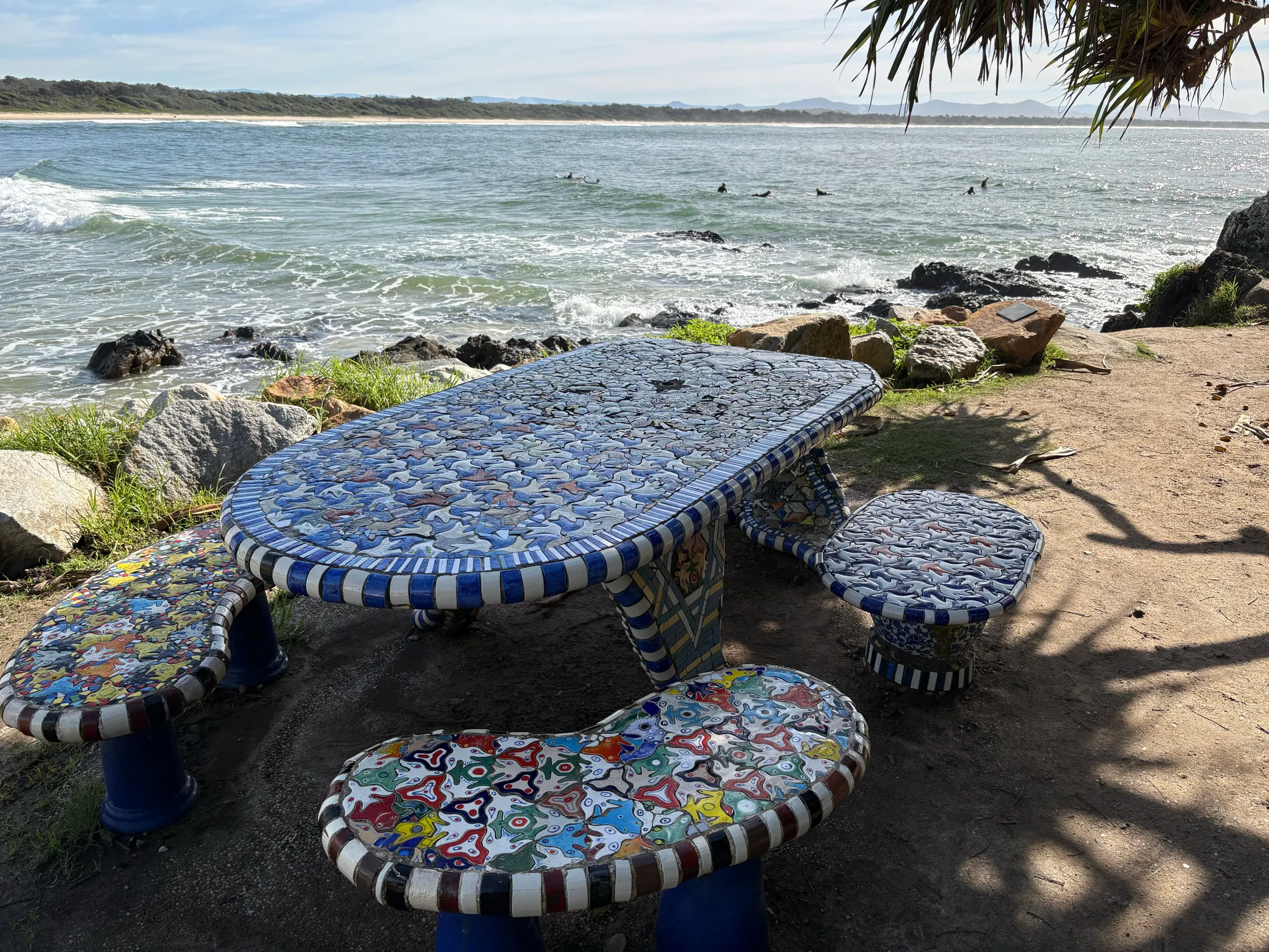 The mosaic Whale Table picnic setting on the Scotts Head foreshore, with surfers in the bay behind
