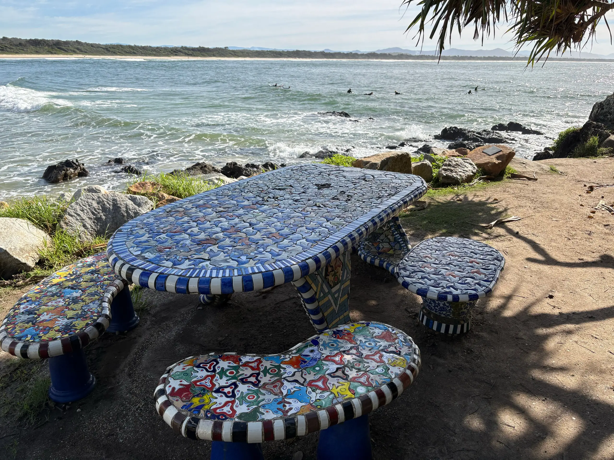 The mosaic Whale Table picnic setting on the Scotts Head foreshore, with surfers in the bay behind