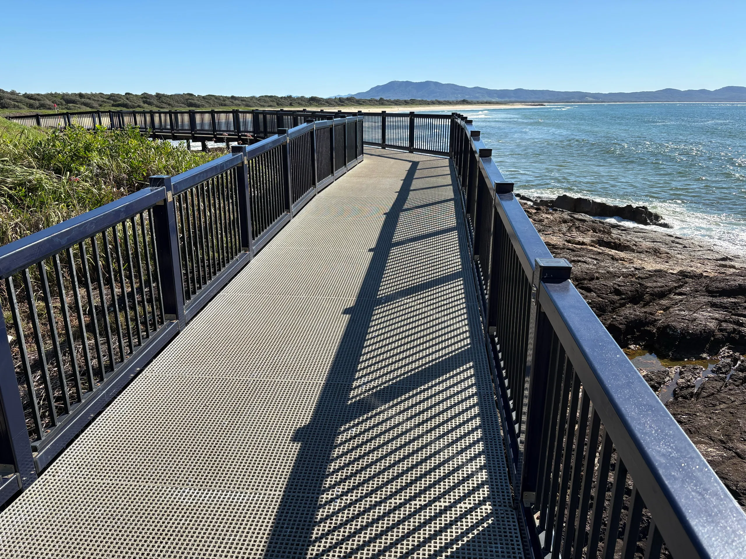Coastal boardwalk leading toward the beach with a mountain backdrop at South West Rocks