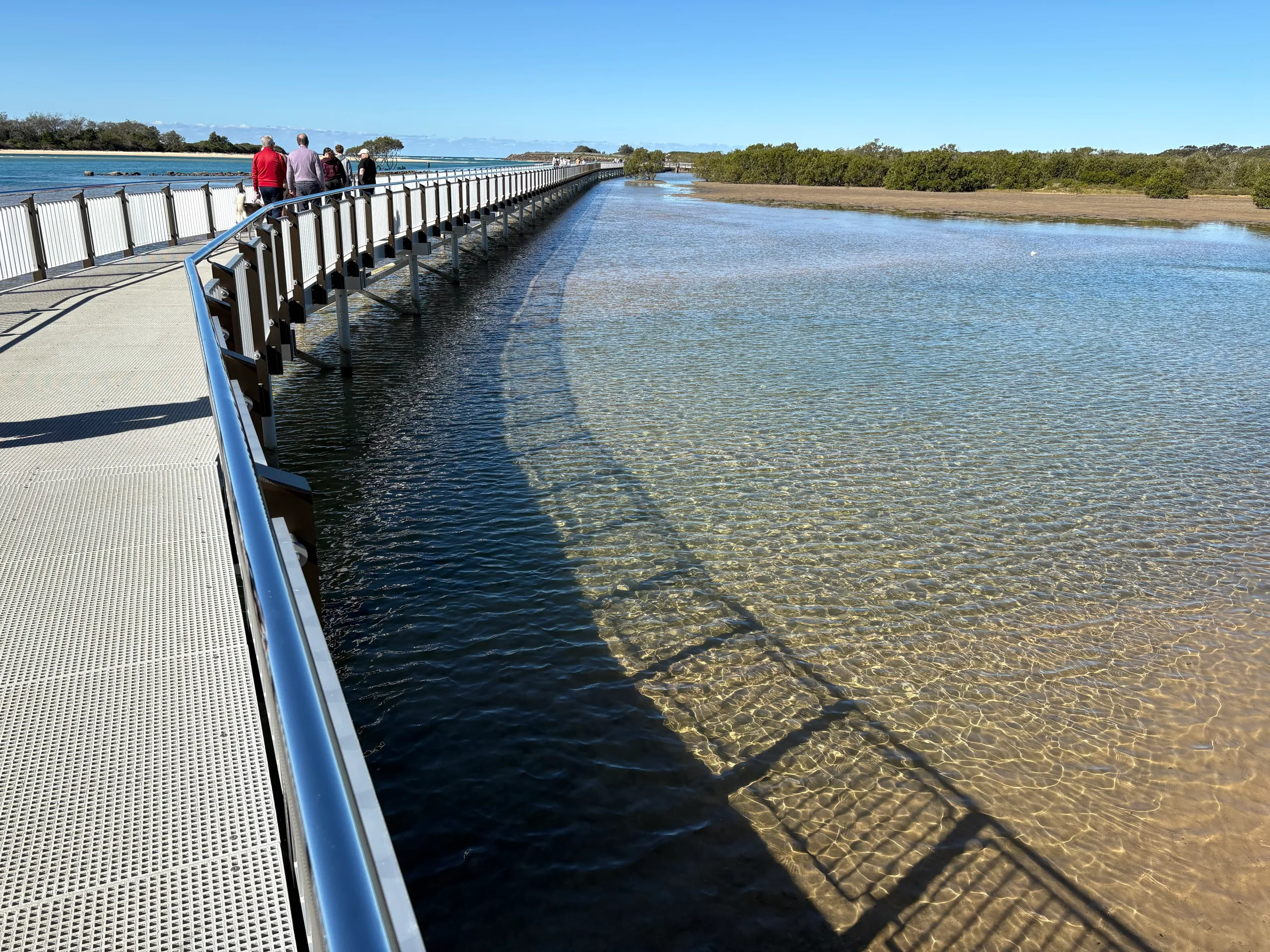 Walkers on the Urunga Boardwalk beside crystal-clear shallow water, looking out toward the ocean