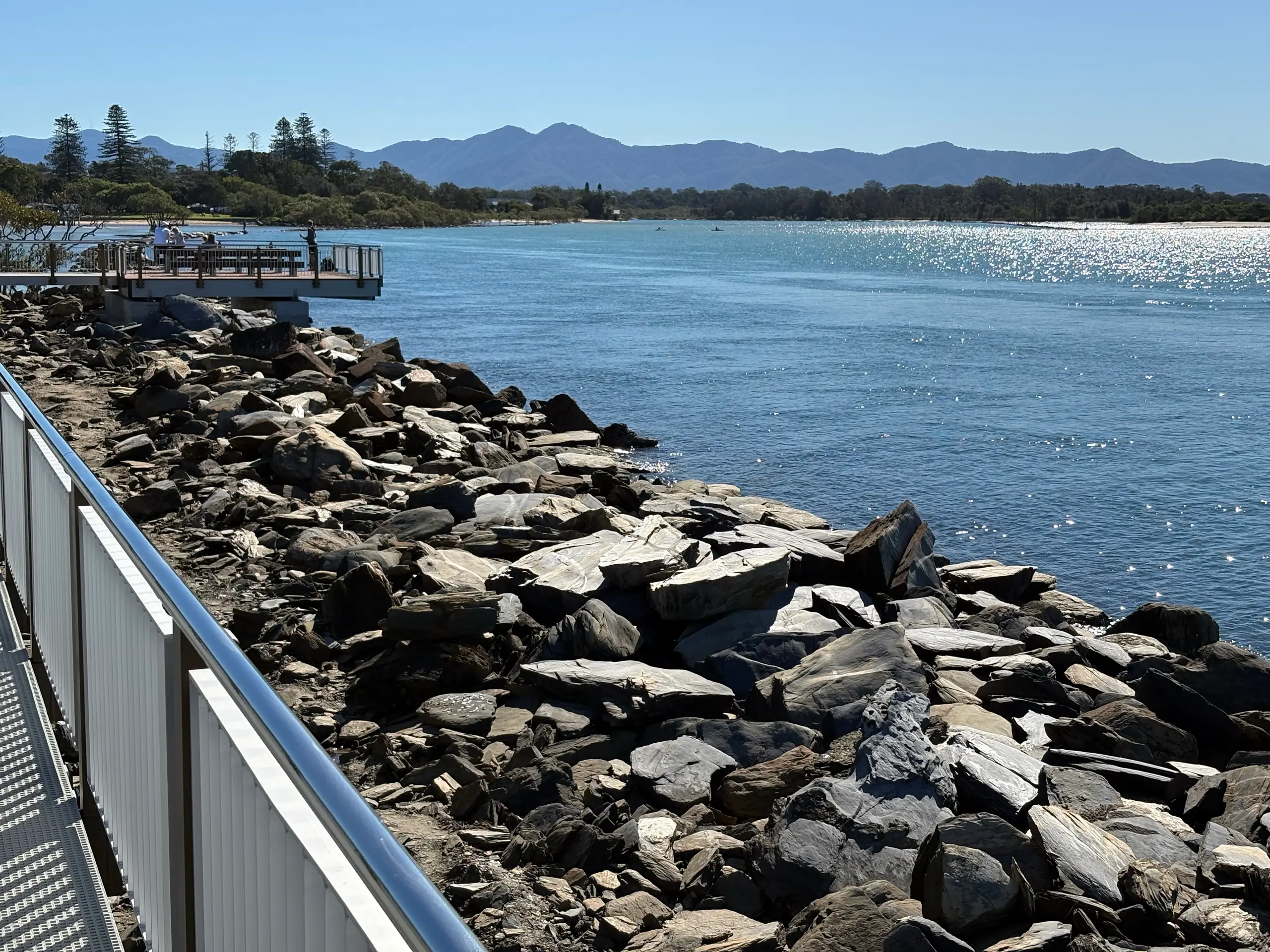 The rock breakwall and river mouth at the end of the Urunga Boardwalk, with mountains in the distance